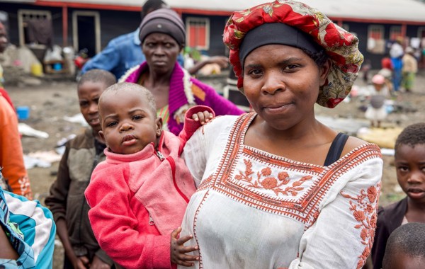 En kvinne med hodeplagg holder et barn på armen. I bakgrunnen ser vi andre mennesker og en skole i DR Kongo. 
