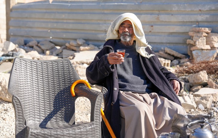 Eldre mann sitter på byggeplass med en kopp te i hånda og tørkle over hodet. 