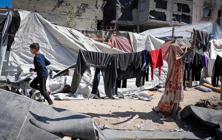 En ung gutt løper på en grusvei omringet av telt og klær på tørkesnorer. I bakgrunnen er det et murhus med store skader. 