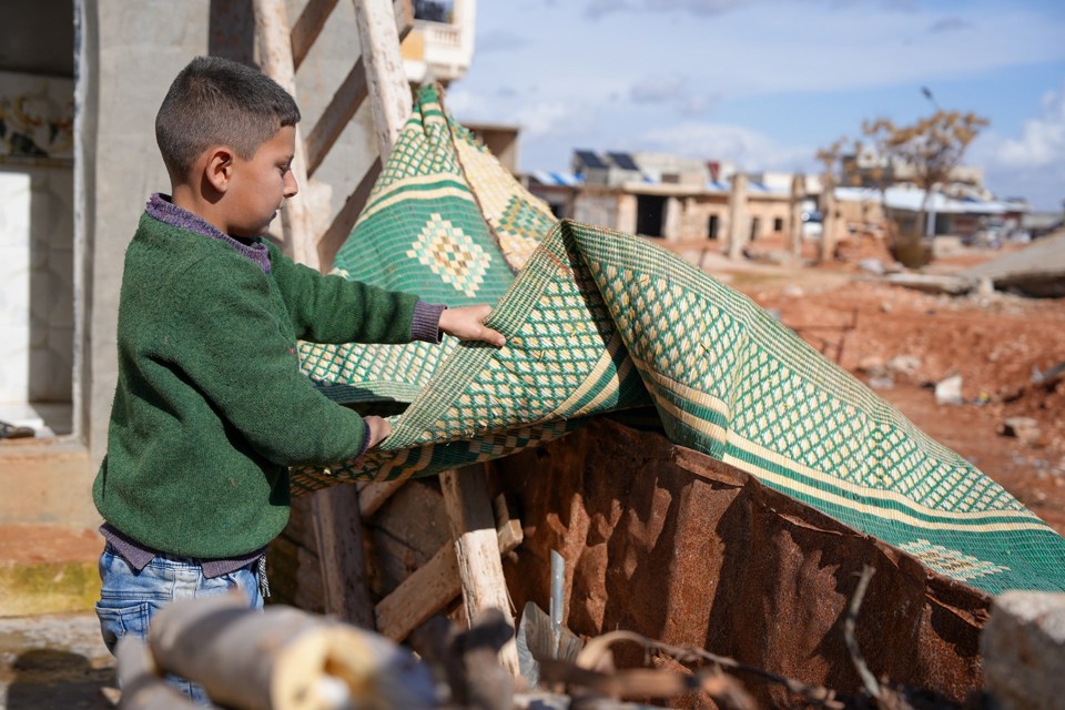 En gutt med grønn genser rister et grønt teppe. Bak er det ruiner. 