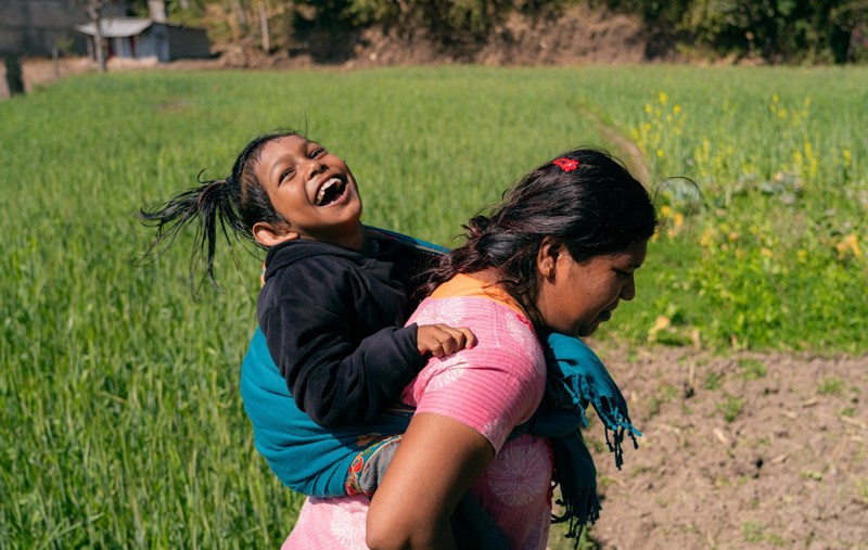 Jente sitter på ryggen til moren sin i en grønn åker i Nepal. Jenta smiler og ler.