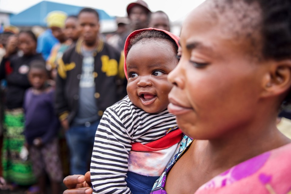 En mor holder et barn som smiler på armen. I bakgrunnen ser vi flere mennesker og et slags telt. 