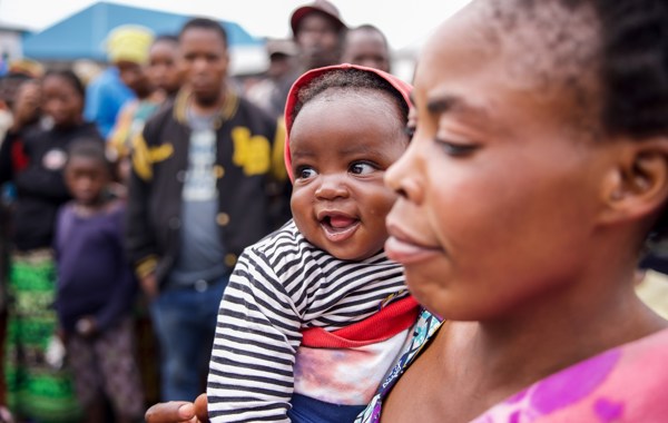 En mor holder et barn som smiler på armen. I bakgrunnen ser vi flere mennesker og et slags telt. 