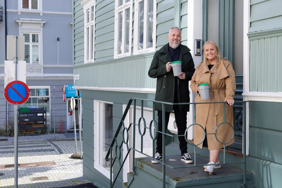 En blond kvinne i beige kåpe og en mann med skjegg og svart frakk står med bøsser utenfor et hus i en by. 