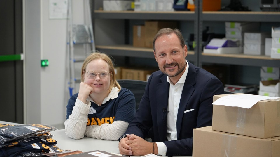 Kronprins Haakon sitter ved siden av ung kvinne med briller på et lagerrom med esker og t-skjorter rundt seg. 