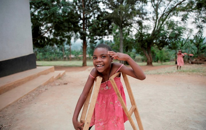 En smilende jente med rosa kjole står ute med krykker og løftet hånd.