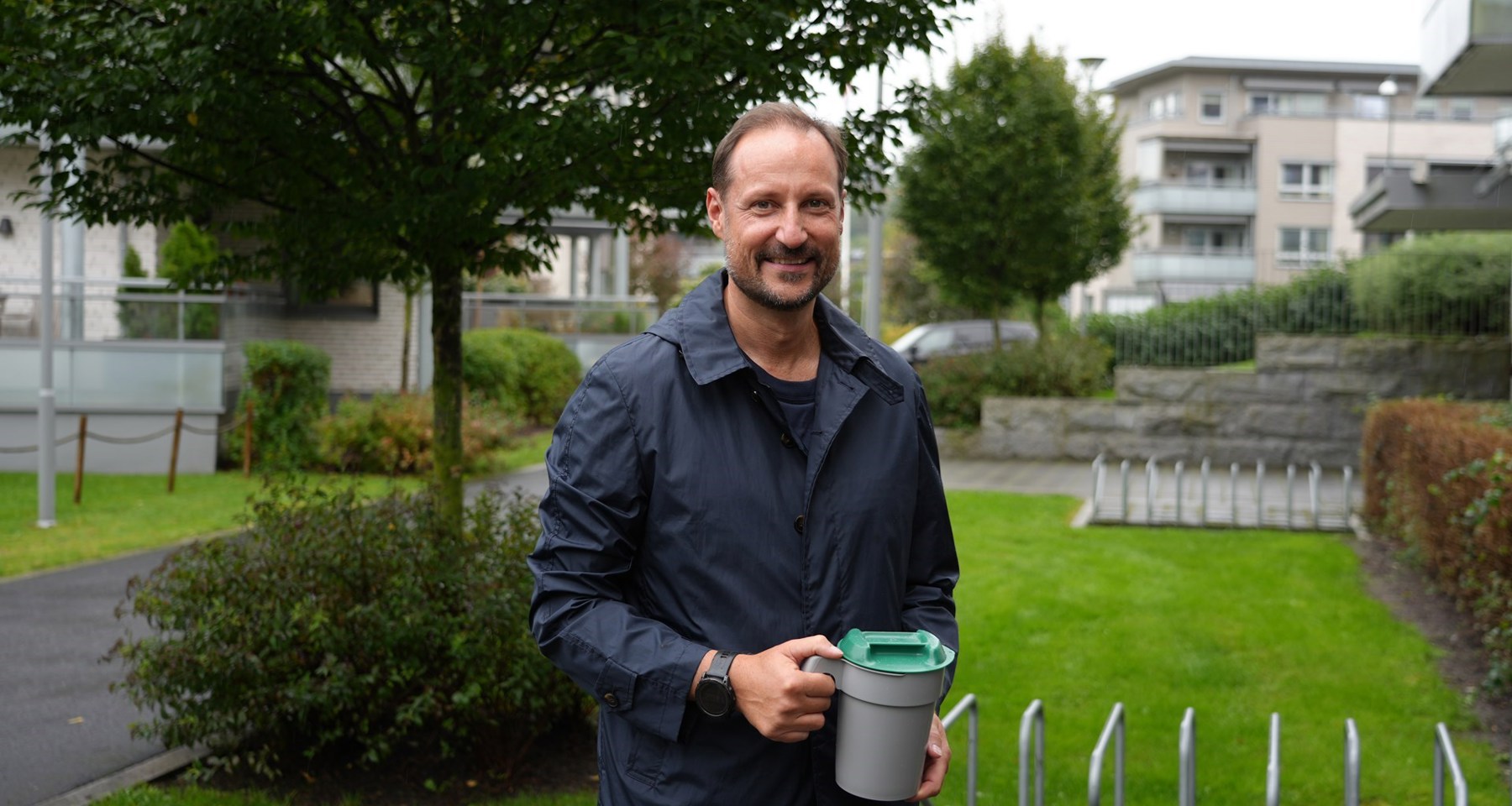 Kronprins Haakon står smilende med blå jakke i et boligområde med en bøsse i hendene. 