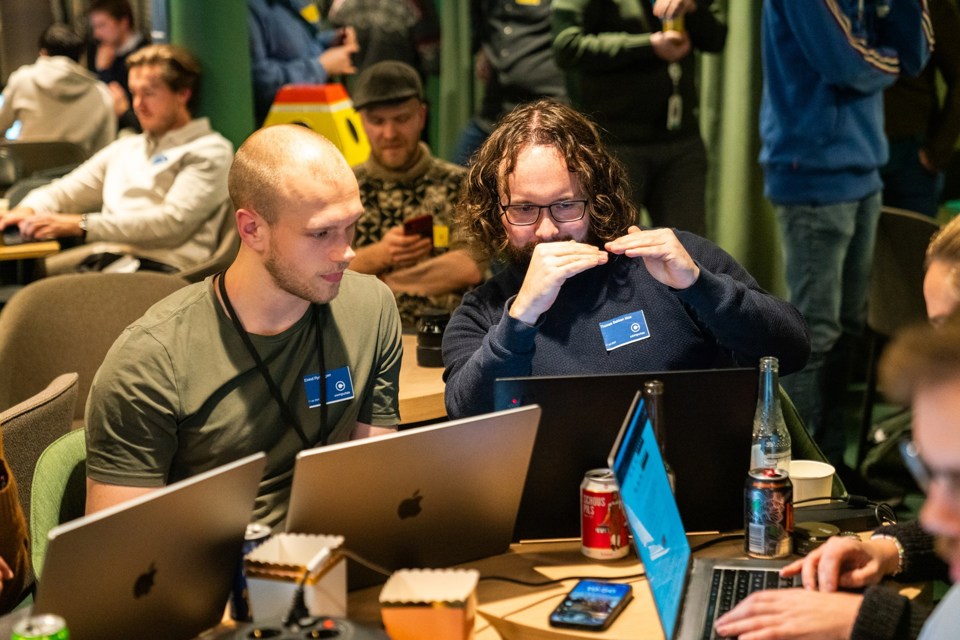To menn sitter foran datamaskiner og snakker sammen.